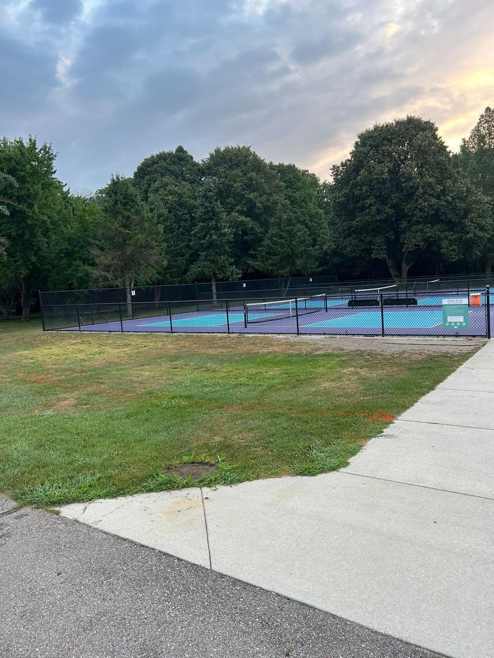Empty outdoor tennis courts surrounded by lush greenery under a cloudy sky.