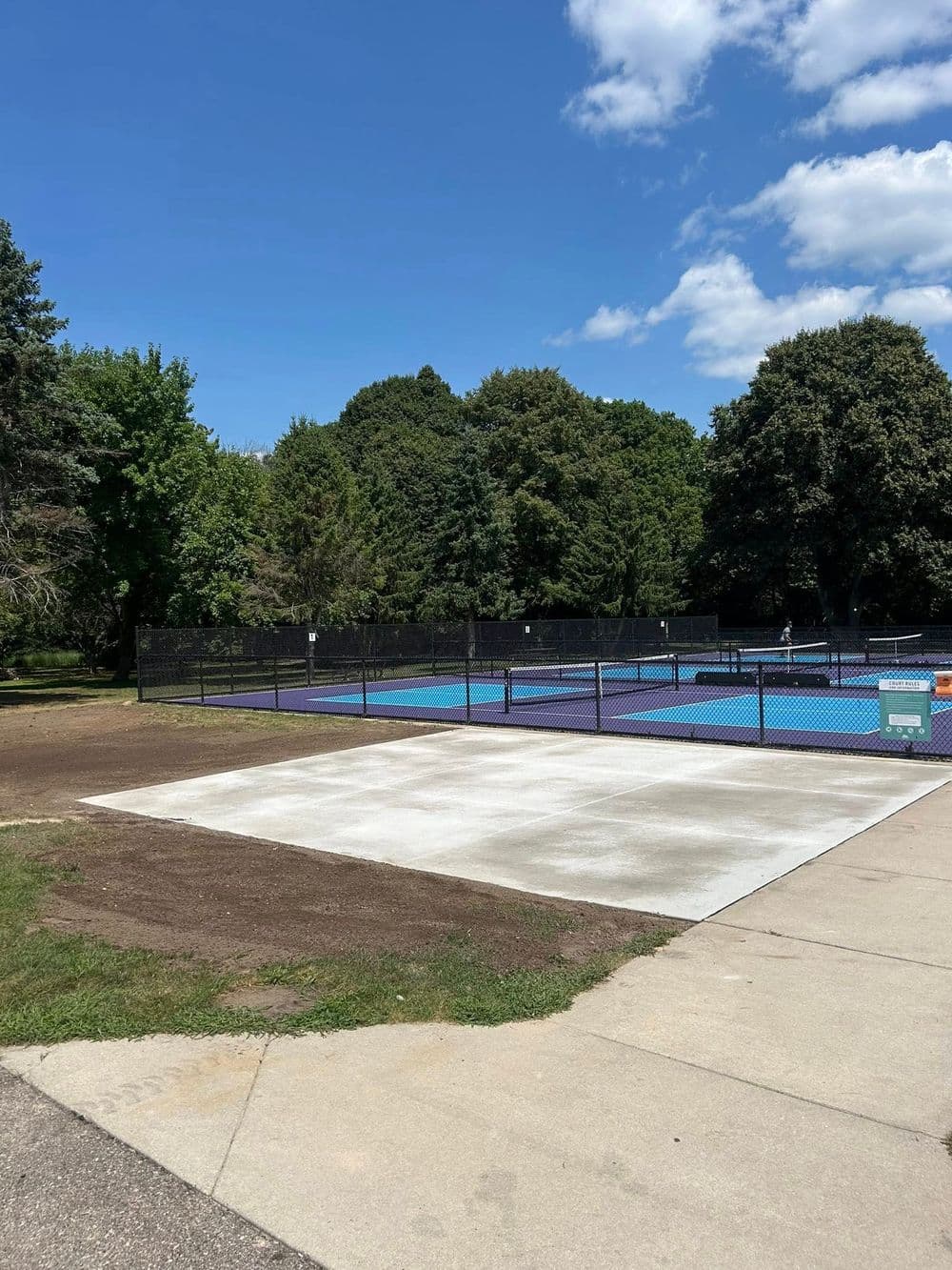 Empty concrete slab near tennis courts in a park surrounded by lush greenery and blue skies.