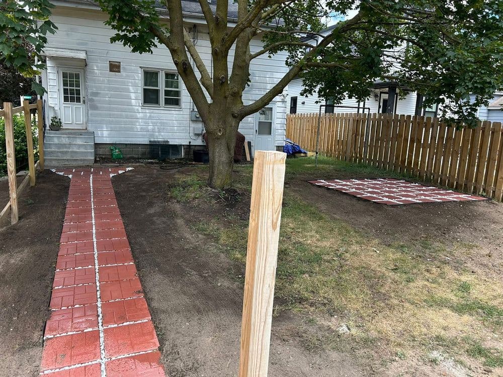 Wooden fence surrounds landscaped yard with a red brick pathway leading to a house.