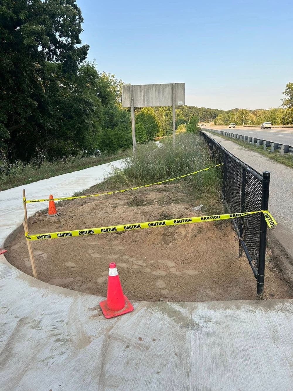 Construction site with caution tape, cones, and a fenced-off area near a highway.
