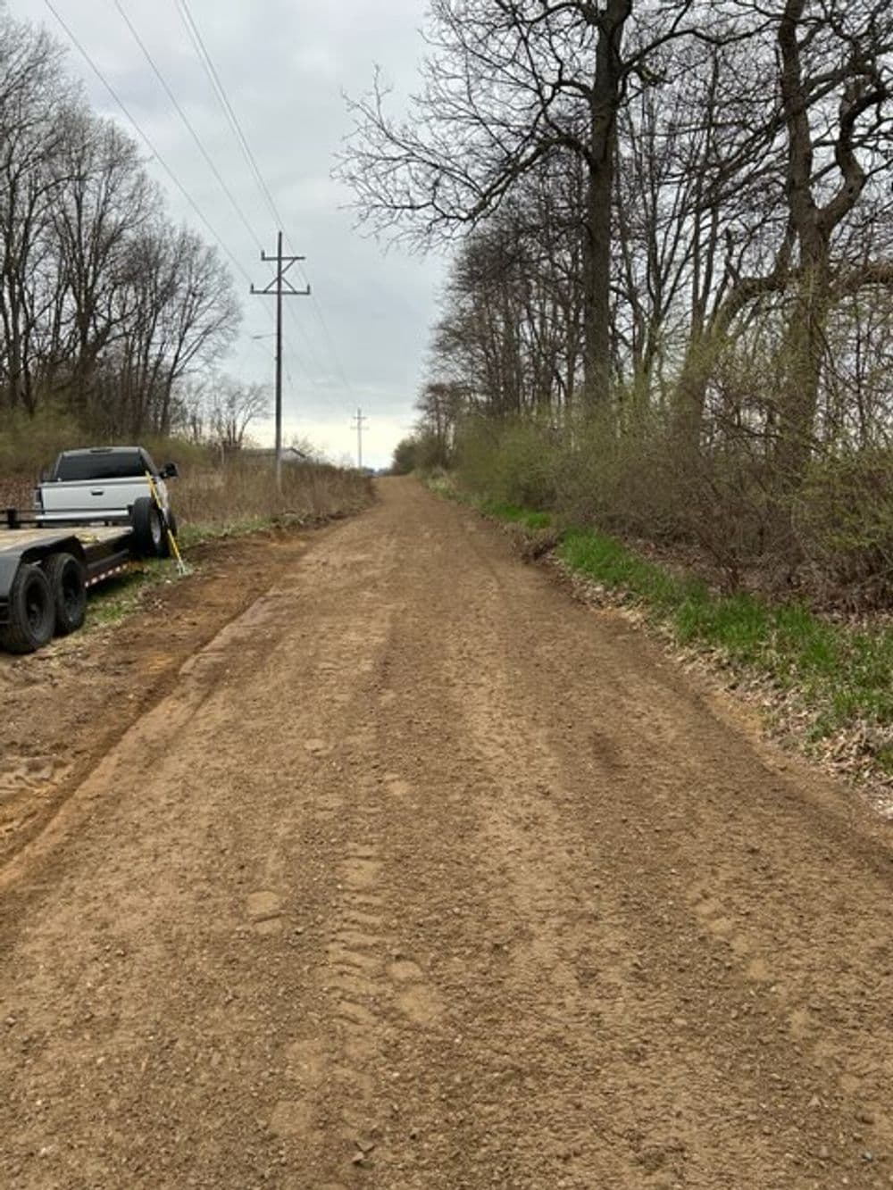Dirt road flanked by sparse trees and power lines, leading into a cloudy horizon.