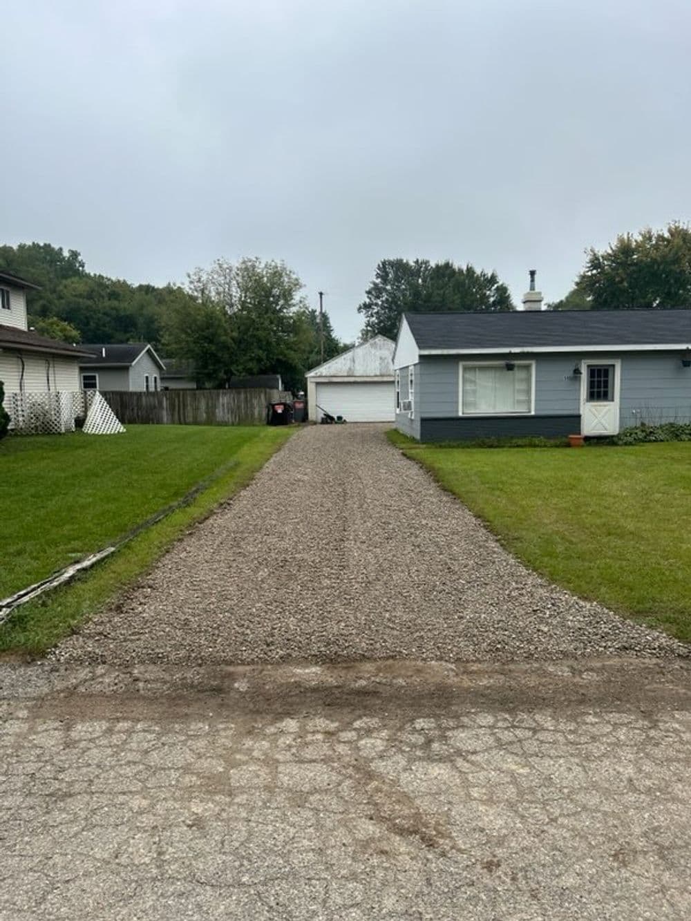 Gravel driveway leading to a garage, surrounded by green grass and houses. Overcast sky.