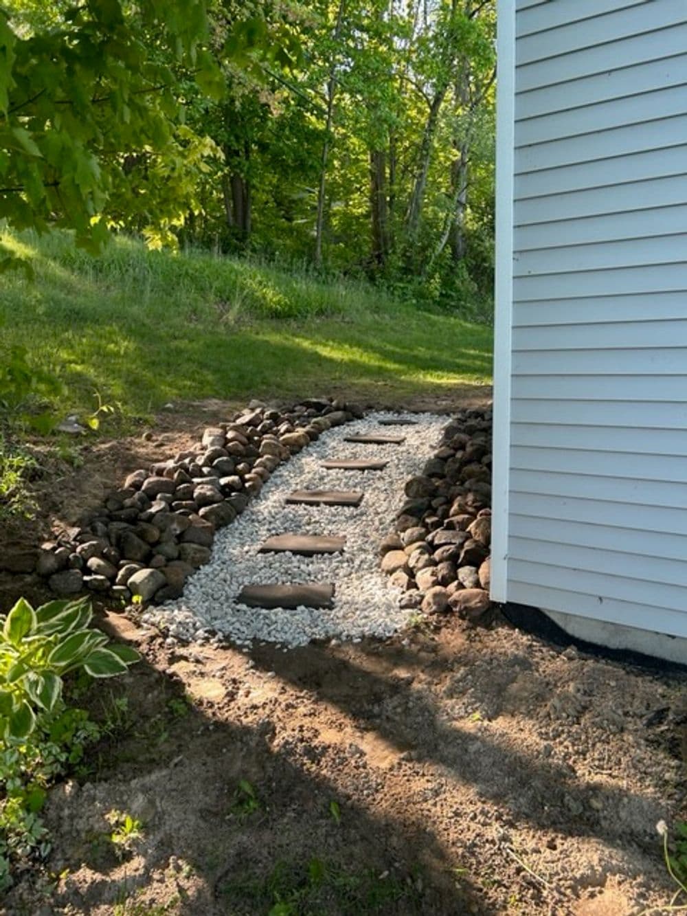 Rock pathway with stepping stones beside a house, surrounded by greenery and gravel.